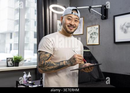 Ein lächelnder asiatischer Tätowierer, der auf Tablet neue Kunstideen ausarbeitet, während er in seinem Tätowierstudio steht Stockfoto