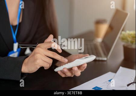 Asiatische Geschäftsfrau hält Stift und benutzt Taschenrechner, während sie im Büro mit Laptop und Dokumenten an ihrem Schreibtisch sitzt Stockfoto
