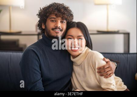 Lächelnder asiatischer Mann und hispanische Frau, die zu Hause auf der Couch sitzt Stockfoto
