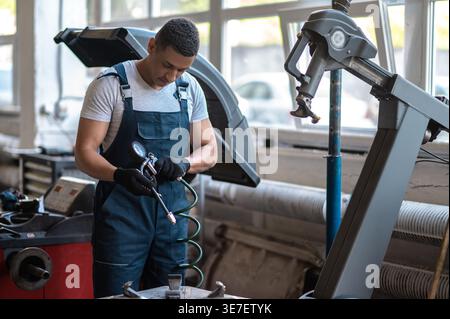Fokussierter afroamerikanischer männlicher Automechaniker mit Handschuhen, der ein kalibriertes Reifendruckmessgerät hält, steht in der Autowerkstatt Stockfoto