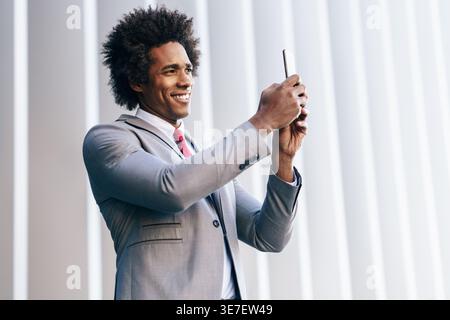 Ein lächelnder schwarzer Geschäftsmann mit Afro-Haaren trägt einen grauen Anzug und rote Krawatte, der Fotos mit dem Smartphone macht, während er im Büro steht Stockfoto