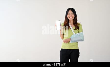 Lächelnde asiatische Frau mit Armschlinge, die den Smartphone-Bildschirm vor grauem Hintergrund zeigt Stockfoto