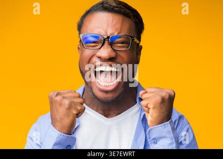 Wütender Schwarzer Mann mit Brille mit geballten Fäusten in der Nähe des Gesichts, der vor gelbem Hintergrund im Studio steht Stockfoto