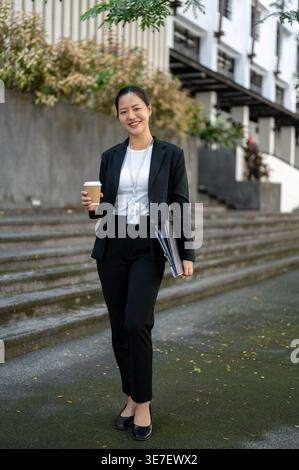 Lächelnde asiatische Geschäftsfrau, die tagsüber eine Tasse Kaffee und Dokumente hält, während sie auf der Straße steht Stockfoto