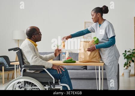 Schwarze Betreuerin, die am Tisch steht und dem schwarzen männlichen Patienten, der zu Hause im Rollstuhl sitzt, eine Flasche Saft schenkt Stockfoto