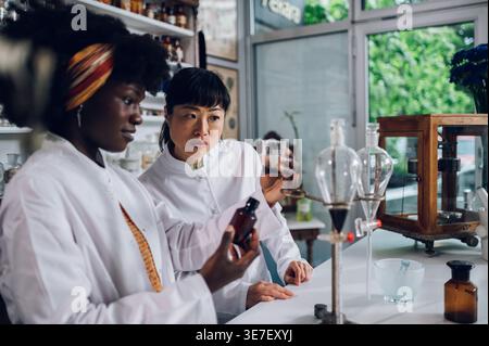 Schwarze Chemikerin hält Becher mit extrahierter Flüssigkeit aus Separatortrichter und Glasflasche, die mit einer japanischen Kollegin in Vintage... Stockfoto
