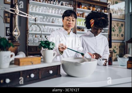 Japanische Apothekerin, die Tee packt, steht mit schwarzer Kollegin, die Medizin in Mörtel in Vintage-Apotheke mischt Stockfoto