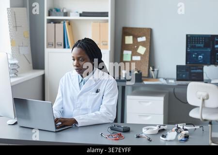 Fokussierte schwarze Computeringenieurin mit Flechten, die einen Labormantel tragen und am Laptop arbeiten, tagsüber im Qualitätskontrolllabor am Tisch sitzen Stockfoto