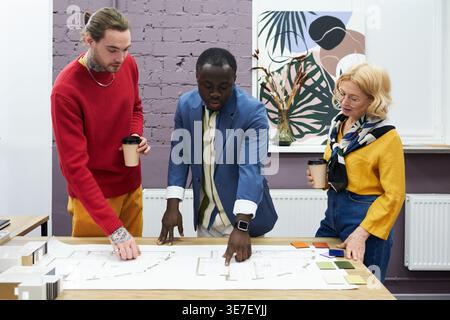 Schwarzer Geschäftsmann, der auf Blueprint zeigt und sich mit Kollegen trifft, die neben ihm am Tisch im Büro stehen Stockfoto