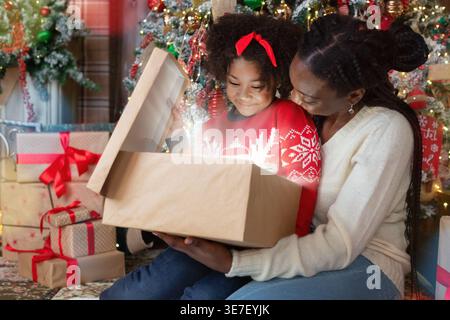 Lächelnde Schwarze Mutter mit Zöpfen und Tochter, die die Geschenkbox öffnet, sitzt dicht zusammen auf dem Teppich in der Nähe des Weihnachtsbaums und Geschenke zu Hause Stockfoto