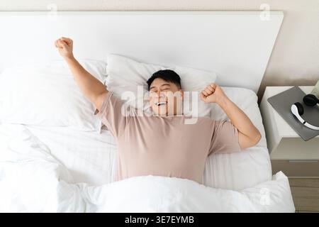 Asiatischer Mann, der die Arme nach dem Aufwachen ausdehnt, während er im Schlafzimmer zu Hause auf dem Bett lag Stockfoto