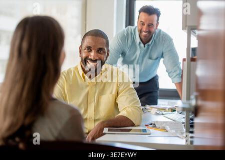 Fröhlicher schwarzer Geschäftsmann sitzt am Tisch mit Dokumenten in der Nähe eines männlichen indischen Kollegen und sieht eine kaukasische Frau im Büro an Stockfoto