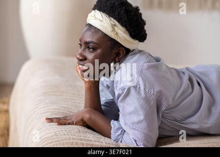 Lächelnde hispanische Frau mit Afro-Tragen Stirnband, das Gesicht auf der Hand liegend, während sie zu Hause auf dem Bett lag Stockfoto