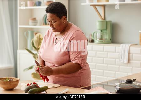 Schwarze Frau mittleren Alters, die Gemüsesalat zubereitete, steht am Tisch in der Küche zu Hause Stockfoto