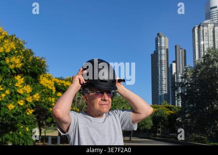 Senior hispanischer Mann mit Sonnenbrille, der Helm steht mit Elektroroller im Stadtpark an sonnigen Tagen Stockfoto
