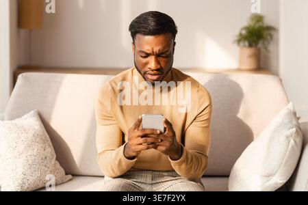 Seriöser Schwarzer Mann, der das Smartphone-Sofa im Wohnzimmer zu Hause benutzt Stockfoto