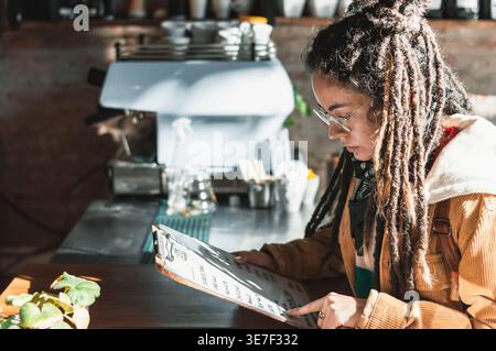 Hispanische Frau mit Dreadlocks, die eine Brille trägt und mit den Fingern auf die Speisekarte zeigt, die tagsüber am Tisch im Café sitzt Stockfoto