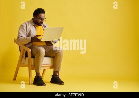 Schockierter asiatischer Mann mit Brille und Blick auf Laptop, der auf Stuhl vor gelbem Hintergrund im Studio sitzt Stockfoto