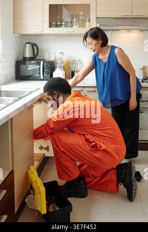 Schwarzer Klempner in orangefarbenem Bodysuit, der Rohr in der Küche installiert, während asiatische Frau den Prozess zu Hause überwacht Stockfoto