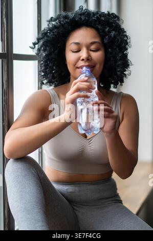 Schwarze Frau mit lockiger Trinkwasserflasche, während sie drinnen auf der Fensterbank sitzt Stockfoto