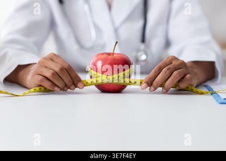 Zugeschnittenes Bild eines schwarzen weiblichen Diätetikers, der roten Apfel mit Bandmaß misst, während er in der Klinik am Tisch sitzt Stockfoto