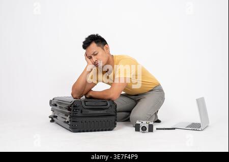 Trauriger asiatischer Mann, der sich auf dem Boden vor weißem Hintergrund im Studio auf das Gepäck gestützt hat Stockfoto