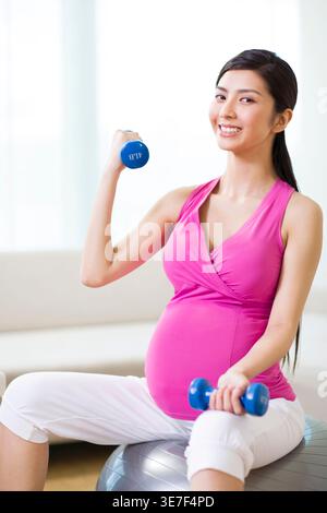 Chinesische schwangere Frau, die mit Hantel zu Hause auf Fitnessball sitzt Stockfoto