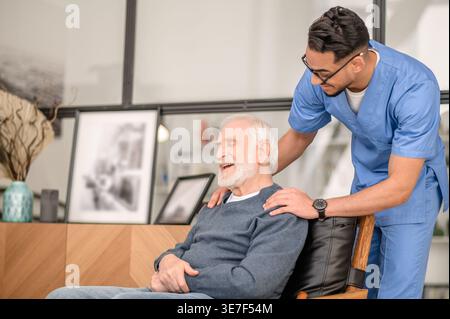 Lächelnder arabischer männlicher Pfleger in Brille, der stehend auf dem Sofa zu Hause sitzt und einen älteren Mann von hinten ansieht Stockfoto