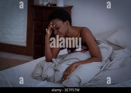 Schwarze Frau mit Hand auf dem Kopf, die an Kopfschmerzen leidet, sitzend auf dem Bett im Schlafzimmer zu Hause Stockfoto