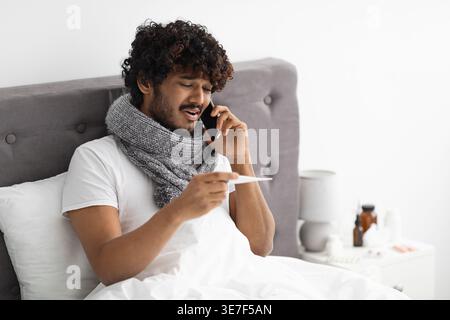 Kranker asiatischer Mann mit lockigen Haaren, der einen Schal um den Hals trägt, Telefonanruf hält und auf das Thermometer blickt, während er auf dem Bett im Schlafzimmer sitzt... Stockfoto