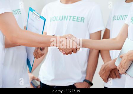 Vietnamesischer Mann, der das Klemmbrett hält, schüttelt Hand mit einem neuen Mitglied der Organisation, während er mit anderen Freiwilligen drinnen steht Stockfoto