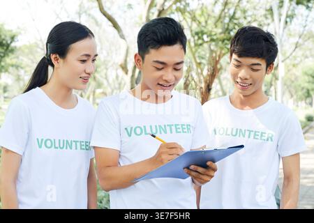 Vietnamesischer Mann mit weißem Hemd, der Namen neuer Freiwilliger in der Liste schreibt, während er am sonnigen Tag zusammen im Park steht Stockfoto