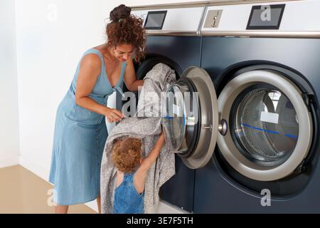 Hispanische Mutter lädt eine Waschmaschine mit Sohn, der ihr hilft, während er in der Wäscherei steht Stockfoto