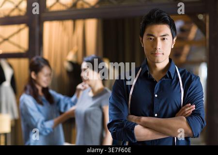 Lächelnder chinesischer Modedesigner mit Maßband um den Hals, der mit überkreuzten Armen im Geschäft steht Stockfoto