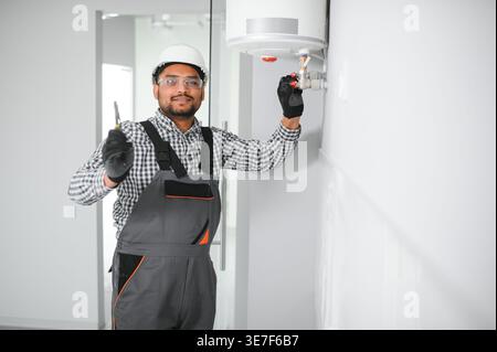 Indischer Klempner mit Uniform und Schutzhelm, der im Bad an der Heizung steht und arbeitet Stockfoto