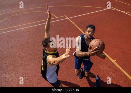 Schwarzer männlicher Athlet, der am sonnigen Tag Baskteball spielt, mit männlichem Teamkollegen aus gemischten Rassen, der ihn während des Trainings auf dem Platz im Freien blockiert Stockfoto