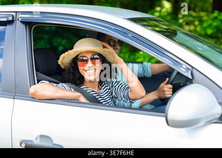 Lächelnde Frau aus dem Nahen Osten mit lockigem Haar, Hut und Sonnenbrille, die bei Sonnenschein mit ihrem Freund im Auto fährt Stockfoto