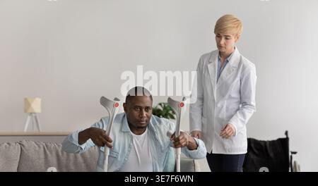 Behinderter Schwarzer Mann mit Krücken, der vom Sofa aufsteht und von einer kaukasischen Therapeutin während der Rehabilitation in der Klinik unterstützt wird Stockfoto