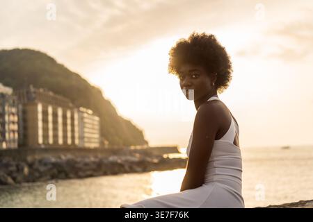Schwarze Frau mit Afro-Frisur, die tagsüber auf einem Betonzaun an der Strandpromenade sitzt Stockfoto