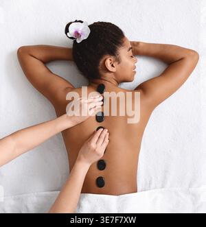 Schwarze Frau mit geschlossenen Augen und Kopf auf einem Handtuch liegend mit dem Gesicht nach unten auf dem Massagetisch, während die Therapeutin in der Kurklinik heiße Steine auf den Rücken legt Stockfoto