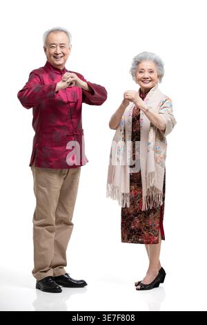 Lächelndes chinesisches Seniorpaar in traditioneller Kleidung, das Faust und Palmensalut macht, während es vor weißem Hintergrund steht Stockfoto