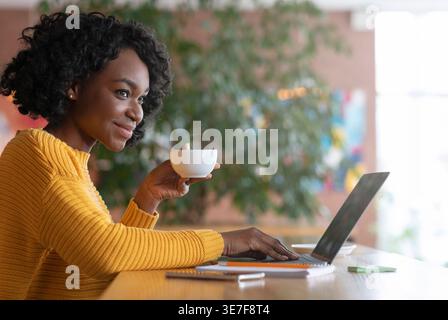Schwarze Frau trinkt Kaffee, während sie am Laptop arbeitet und am Tisch im Café sitzt Stockfoto