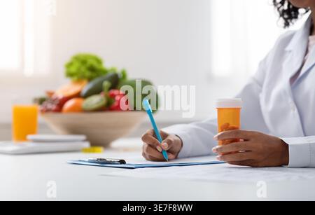 Zugeschnittenes Bild einer schwarzen Ernährungsberaterin, die eine Flasche Pillen hält und auf das Klemmbrett schreibt, während sie mit einer Schale Obst und Gemüse am Tisch sitzt... Stockfoto