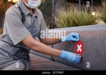 Gemischte Kellnerin mit Gesichtsmaske, Schürze und Handschuhen, die soziale Distanzierung auf dem Sitz anbringen, während sie während des Aufenthalts auf dem Sofa im Café sitzt... Stockfoto