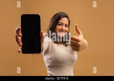 Begeisterte arabische Frau, die ein riesiges Smartphone mit leerem Bildschirm und Daumen nach oben zeigt, die neueste mobile App oder coole Website vorführt, Mockup, beigefarbener Hintergrund, Stockfoto