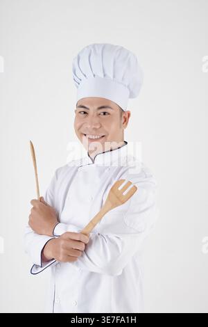 Asiatischer männlicher Koch mit überkreuzten Armen, der im Studio vor weißem Hintergrund Küchenwerkzeuge aus Holz hält Stockfoto