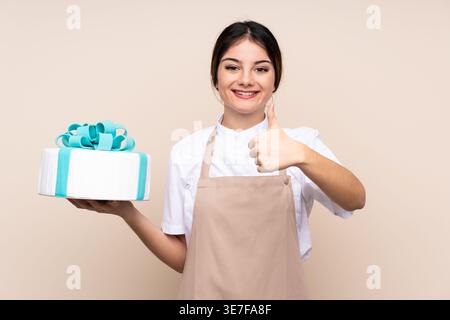 Die hispanische Konditorin zeigt die Daumen hoch und hält im Studio Kuchen vor beigem Hintergrund Stockfoto