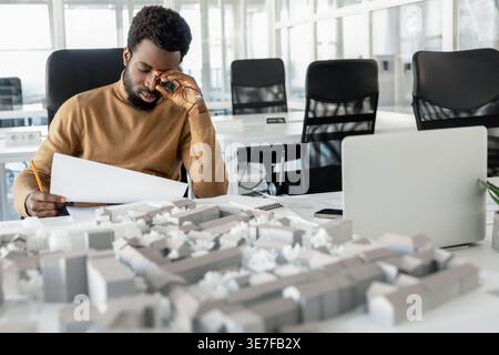 Müder afroamerikanischer Architekt, der Baupläne im Büro überprüft Stockfoto