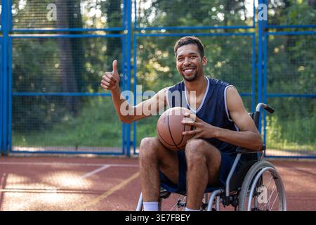 Schwarzer Mann mit Behinderung, der Basketball hält und die Daumen hoch zeigt, während er tagsüber im Rollstuhl auf dem Basketballfeld sitzt Stockfoto