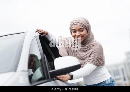 Fröhliche schwarze Frau im Hijab, die tagsüber neben dem Auto in der Stadt steht Stockfoto